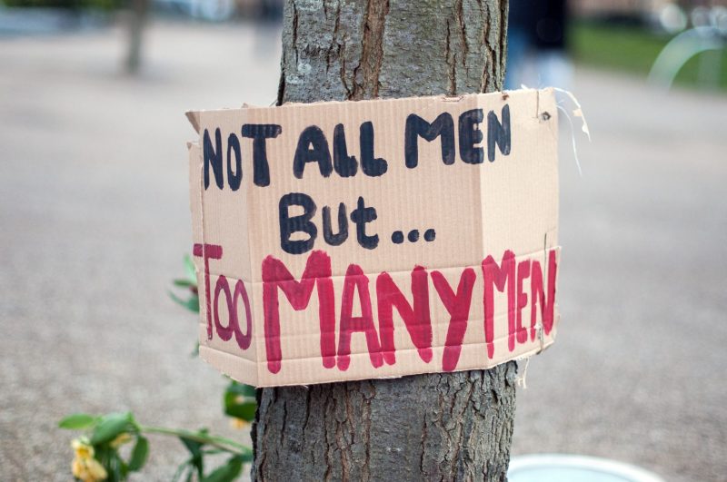A cardboard sign painted saying not all men but too many men hung around a tree