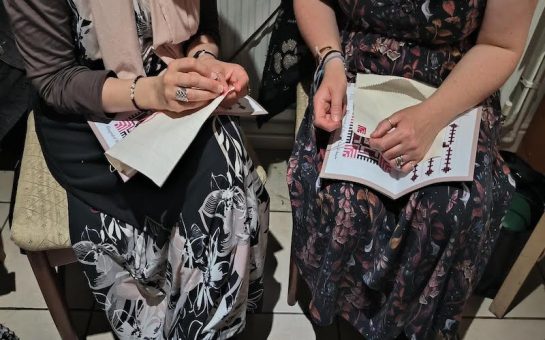Image shows two women sat side by side wearing patterned dresses, stitching on white fabric.