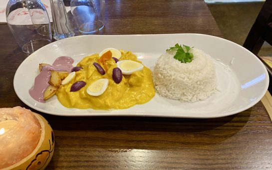 A vibrant yellow chicken curry served with rice, purple olives, and hard-boiled eggs sits on top of a dark wood table at Tierra Peru Islington