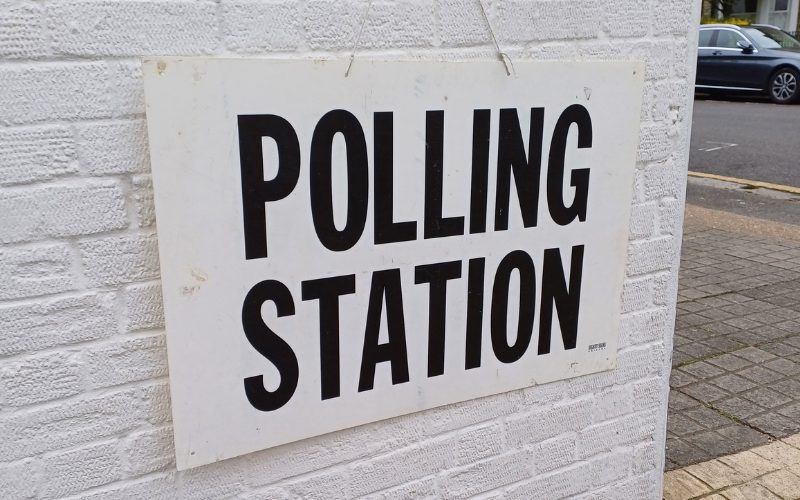 A polling station sign which is hung on a white wall.