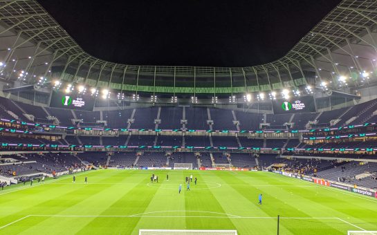 An image of the inside of Tottenham Hotspur stadium, showing the pitch and an empty stand.