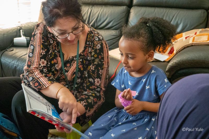 A female charity worker from Doorstep Library reading a book to a small child in a home. 