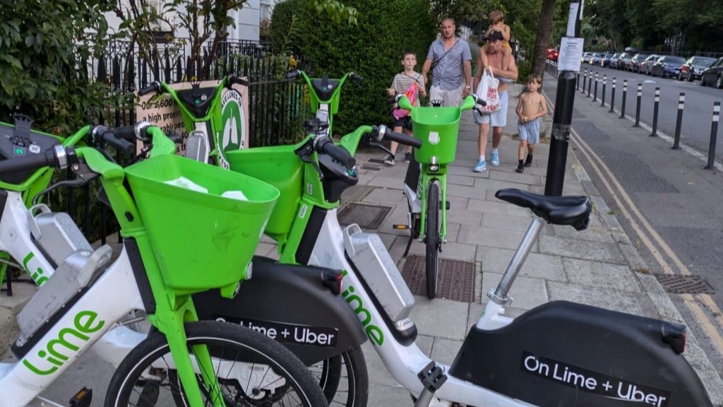 The image shows five Lime bikes parked on the pavement, blocking the path of a family walking on the pavement. The family consists of two men, a child on one's shoulders, and two other young boys.