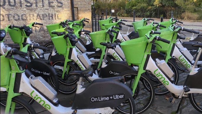 The image shows 16 Lime bikes parked carelessly on a sidewalk, closely grouped together and blocking the pedestrian pathway. The placement of the bikes makes it difficult for people, especially those with mobility issues, to use the sidewalk freely.