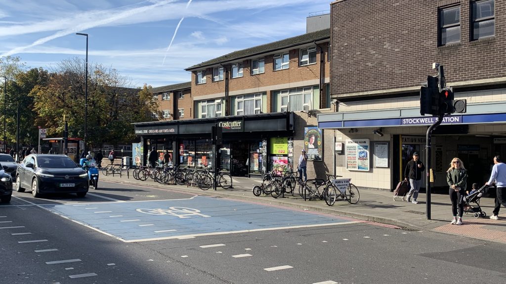 Street view of Stockwell Station in London on a sunny day. The station entrance is on the right, with people walking in and out. Bicycles are parked in front of a Costcutter store next to the station, and several cars and a motorbike are stopped at a traffic light. Trees with autumn leaves are visible in the background.