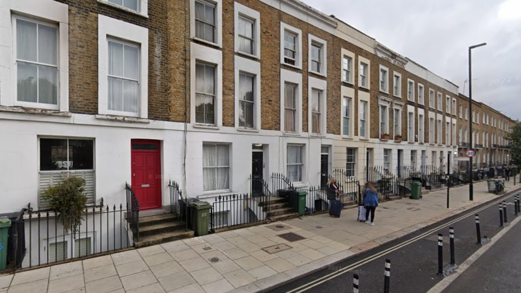 Row of classic terraced houses with brick and white facades on Prince of Wales Road in Chalk Farm, London. A red door stands out among the buildings, and a few people with luggage walk along the pavement. Green bins are lined up by the railings in front of each house.