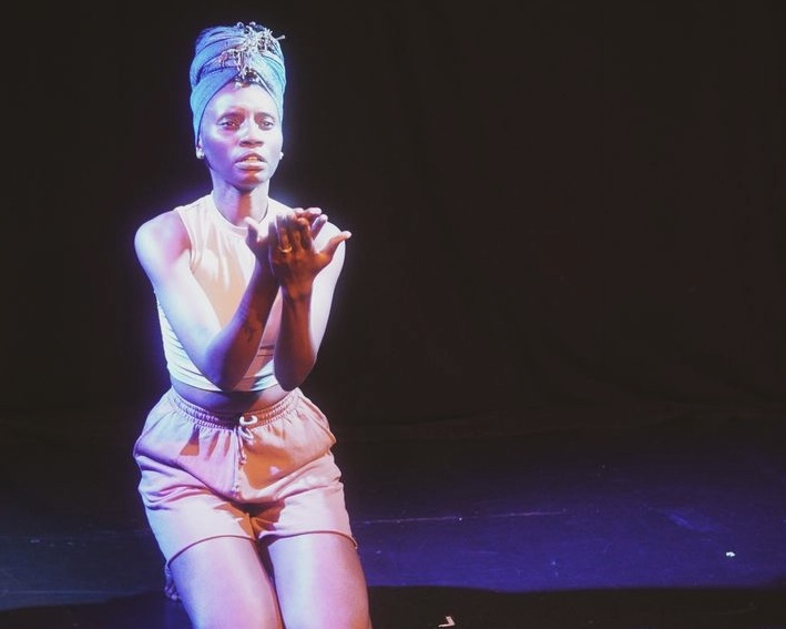 A woman wearing a headwrap, tank top, and shorts performs on stage with a solemn expression, holding her hands out in a gesture of openness. She is lit by a spotlight against a dark background.