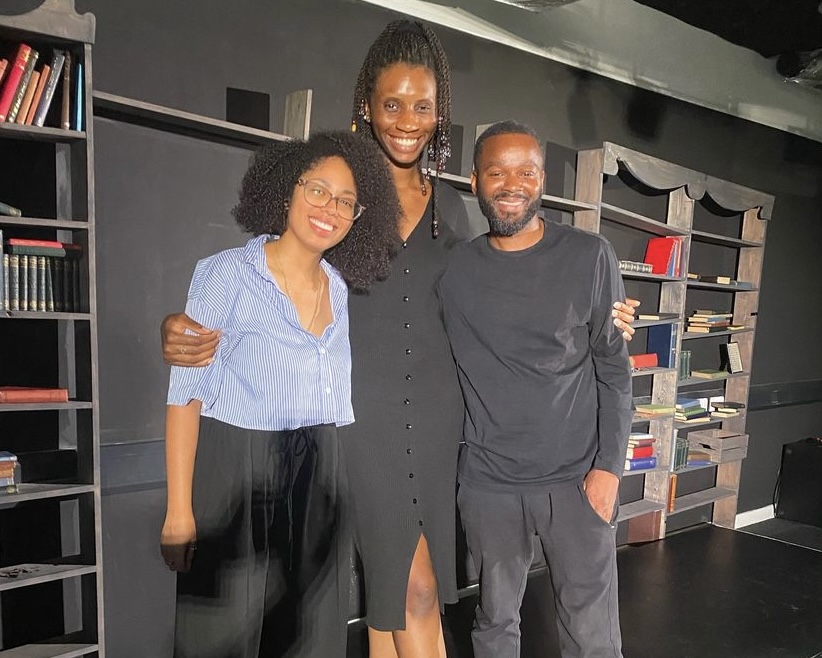 Three people, two women and one man, stand together and smile at the camera on a stage set with bookshelves and props. The woman in the middle is wearing a black dress, while the other two are dressed casually.