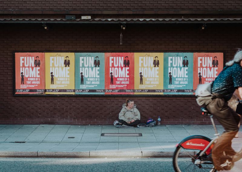 Photo shows a homeless women sitting on the street under posters of 'Fun Home'.