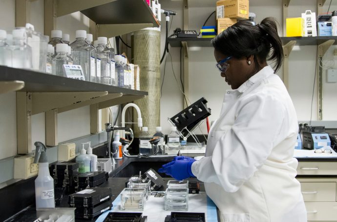 An image of a woman in a white lab technician coat standing over a sink in the lab.