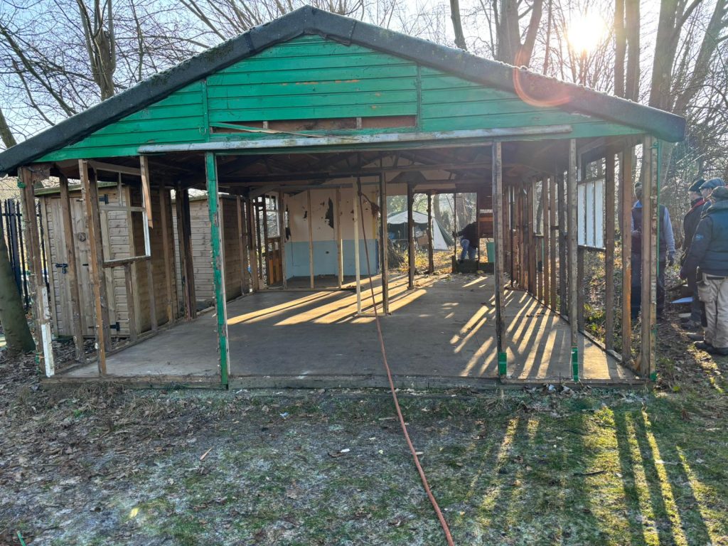 The old hut on site at Waltham Abbey during its disassembly. The sun shines through the bare slats as volunteers gather round. 