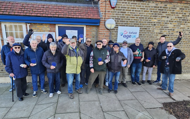 A group of older men smiling outside a building in an ageUK Enfield sign.