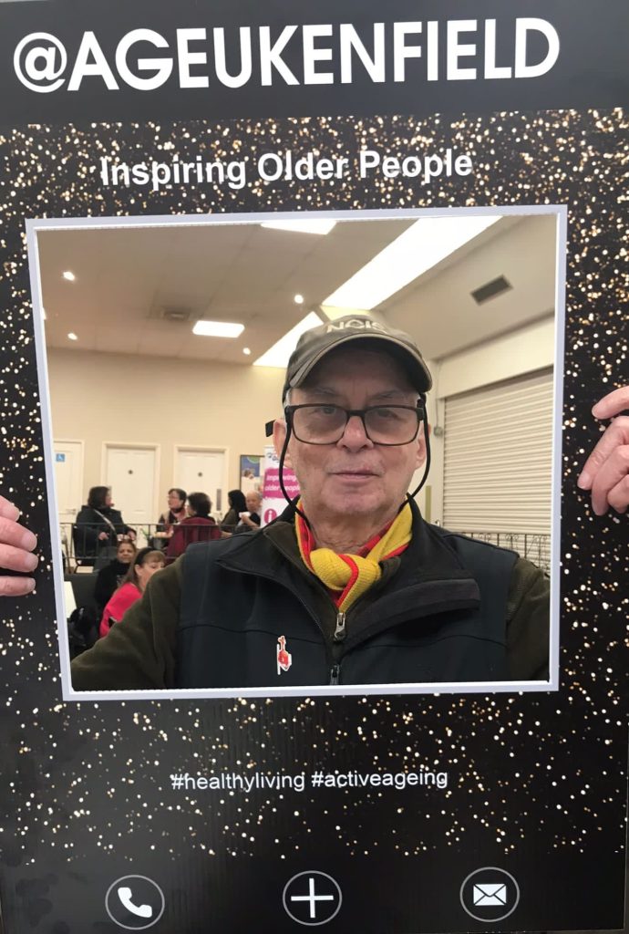 Gordon Tweddle, a older man wearing glasses and a hat posing behind a sign for ageUK Enfield.