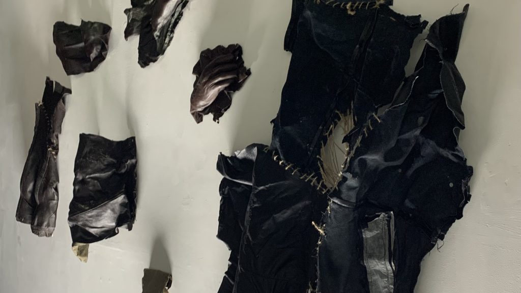 A collection of black leather sculptures is mounted on a light-colored wall. The pieces are irregularly shaped, torn, and stitched together with visible golden-brown thread. Some fragments appear crumpled or folded, while others have jagged edges. The arrangement gives a raw, deconstructed aesthetic, evoking a sense of organic decay or reconstructed garments.