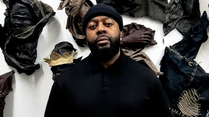 An artist wearing a black beanie and a plain black shirt stands confidently in front of a wall covered with dark, textured leather pieces. The leather is arranged in varied shapes and sizes, its tactile quality giving the wall a raw, yet sophisticated look. The lighting accentuates the rich textures of the leather, casting shadows that enhance the depth and complexity of the installation. The artist’s expression is focused and intentional, clearly engaged in their own creation, as they stand before their work at the exhibition, embodying both the creator and the artwork in a moment of quiet reflection.